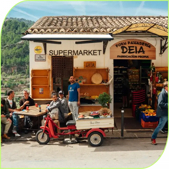 2B LOCAL app Outdoor market stall selling fruits and vegetables, several people seated at a wooden table, a red scooter in front, hills in the background.